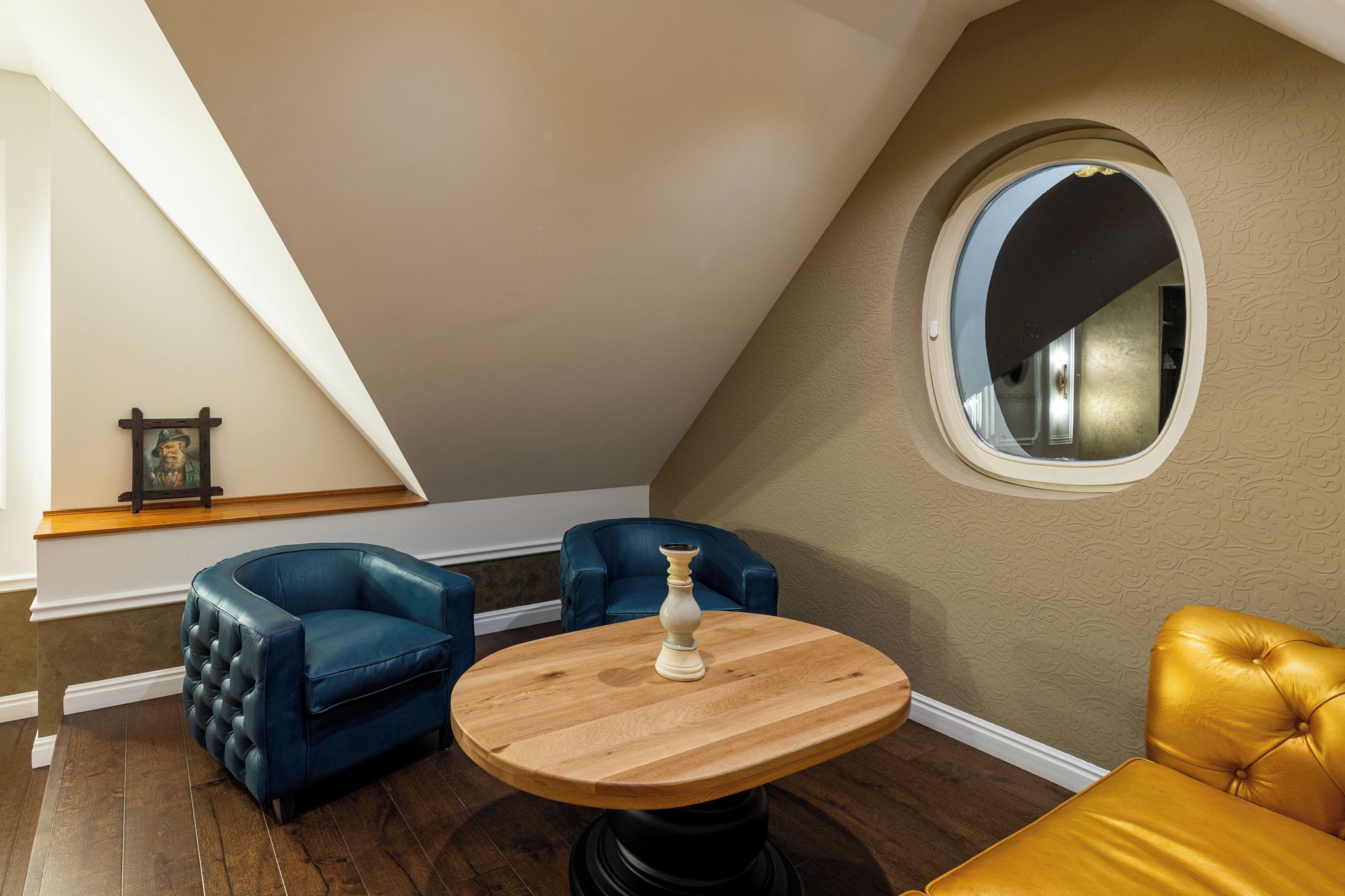 Ein Zimmer mit schrägverdeckter Decke, einem runden Holztisch, zwei blauen Ledersesseln, einem gelben Ledersofa und einem dekorativen Spiegel.
