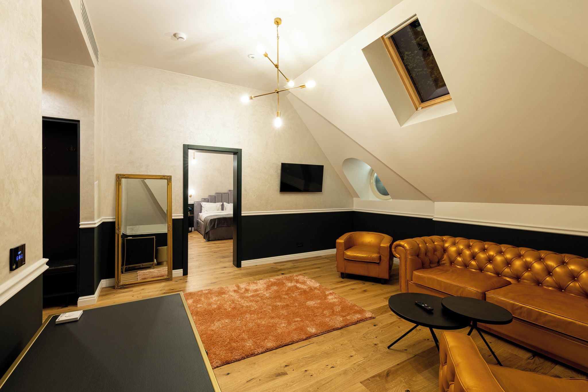 Amplio dormitorio tipo loft con techo alto, techo inclinado con tragaluz, cama grande, sofá de cuero y una mesita pequeña.