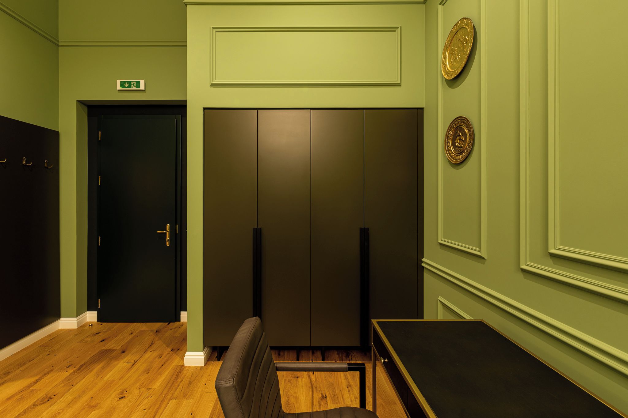 Una habitación con suelo de madera, una puerta negra, un armario negro, un sillón de cuero marrón y una pequeña mesa.