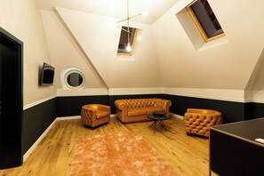 Una habitación en el ático con techo inclinado, un tragaluz redondo, un televisor negro, un espejo redondo, un sofá capitonado, una pequeña mesa redonda y piso de madera.