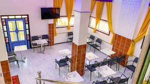 A hotel lobby with a blue door, multiple tables and chairs, a flat-screen TV, and yellow curtains.