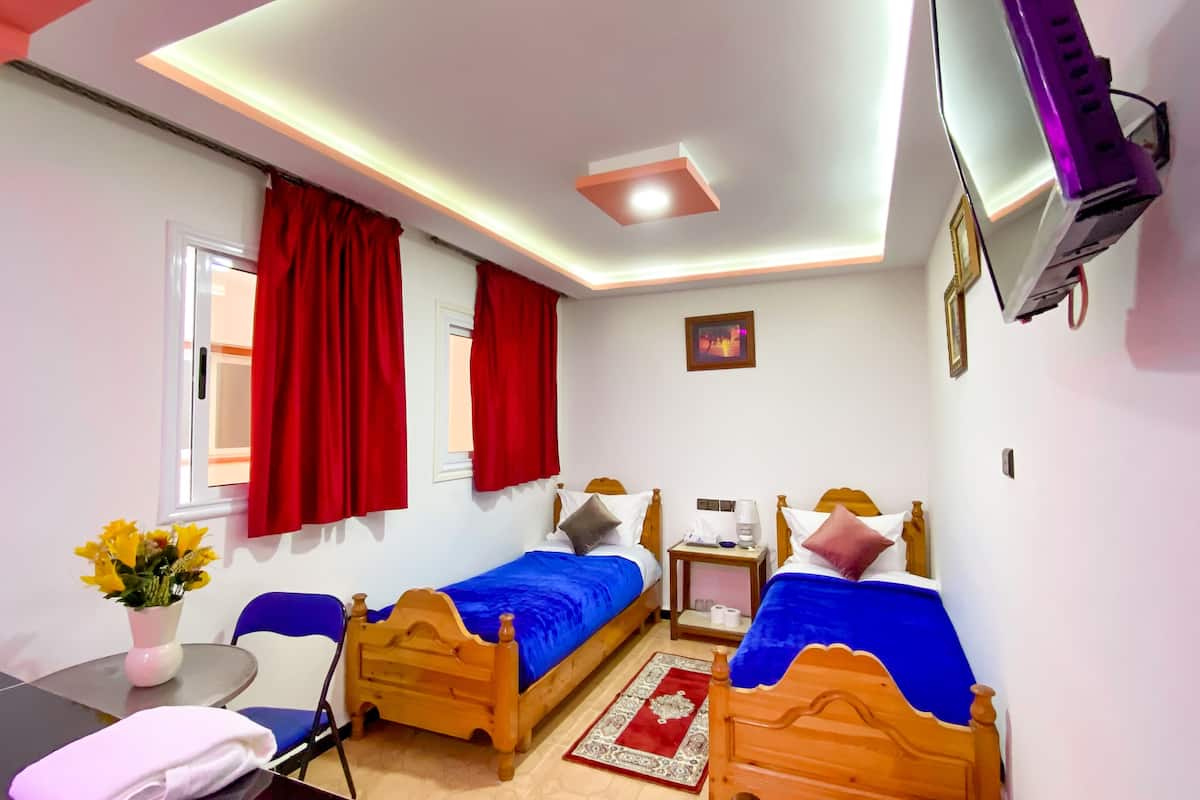 Habitación de hotel con dos camas de madera, un escritorio, una silla, un televisor y un ventanal con cortinas rojas.