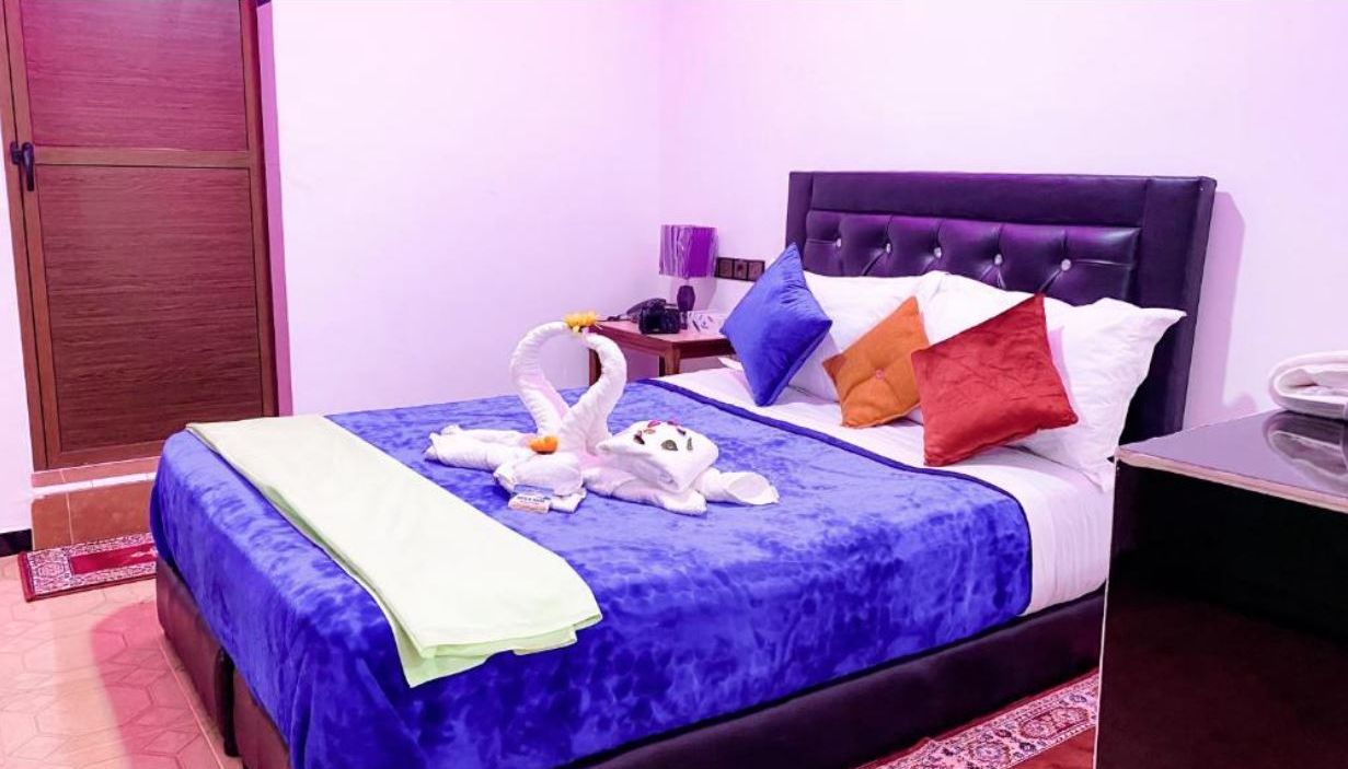 Una cama con colcha azul, cabecera y varios almohadones de distintos colores.