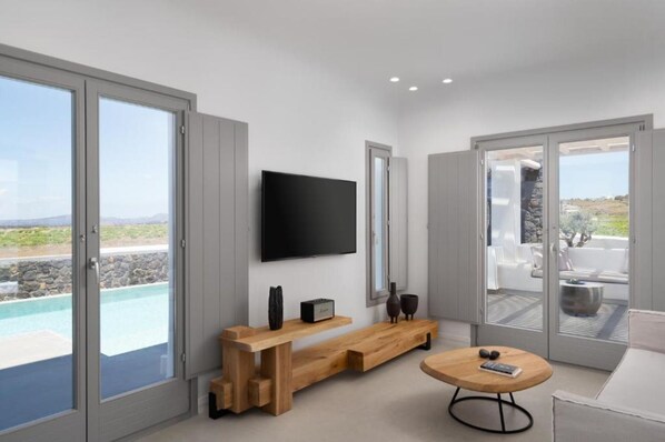 A modern living room with a TV, wooden bench, and round table, large sliding glass doors, and a view of a pool and landscape. - La Ponta Villas&suites (Santorini)