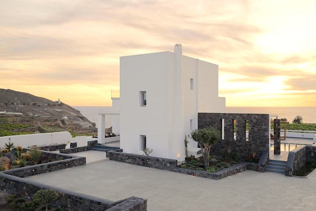 A modern white building with a flat roof, stone accents, and a view of the sea at sunset.