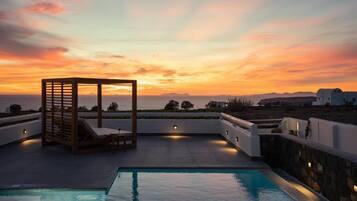 Una piscina en la azotea con una pérgola de madera y sillones de descanso, con vistas a un atardecer y una lejana vista del mar y las colinas.