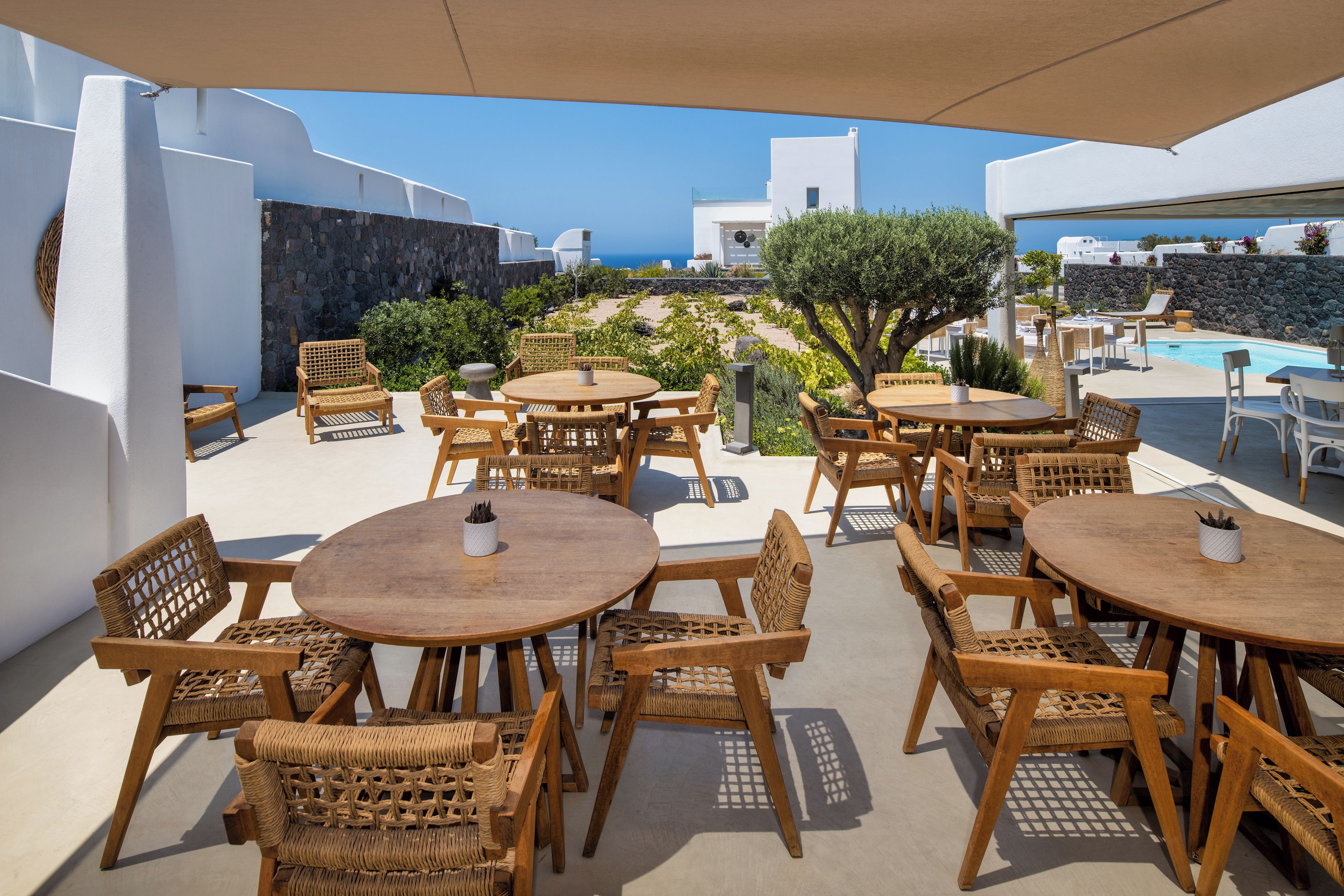 Un área de estar al aire libre con mesas de madera redondas y sillas de mimbre, con vista a una piscina y al mar.