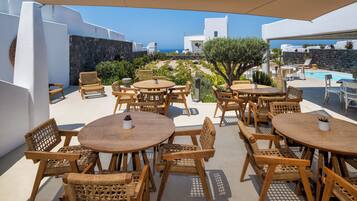 Un área de estar al aire libre con mesas de madera redondas y sillas de mimbre, con vista a una piscina y al mar.