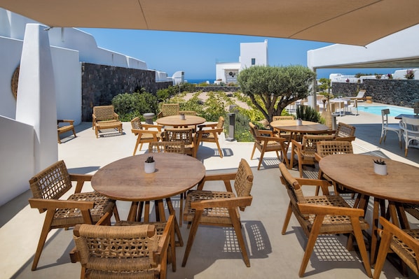 An outdoor seating area with round wooden tables and wicker chairs, overlooking a pool and a view of the sea. - La Ponta Villas&suites (Santorini)