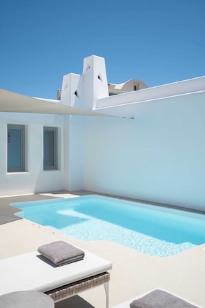 A modern pool area with a clear blue sky, a white building with a unique architectural design, and a lounge chair with a towel. - La Ponta Villas&suites (Santorini)