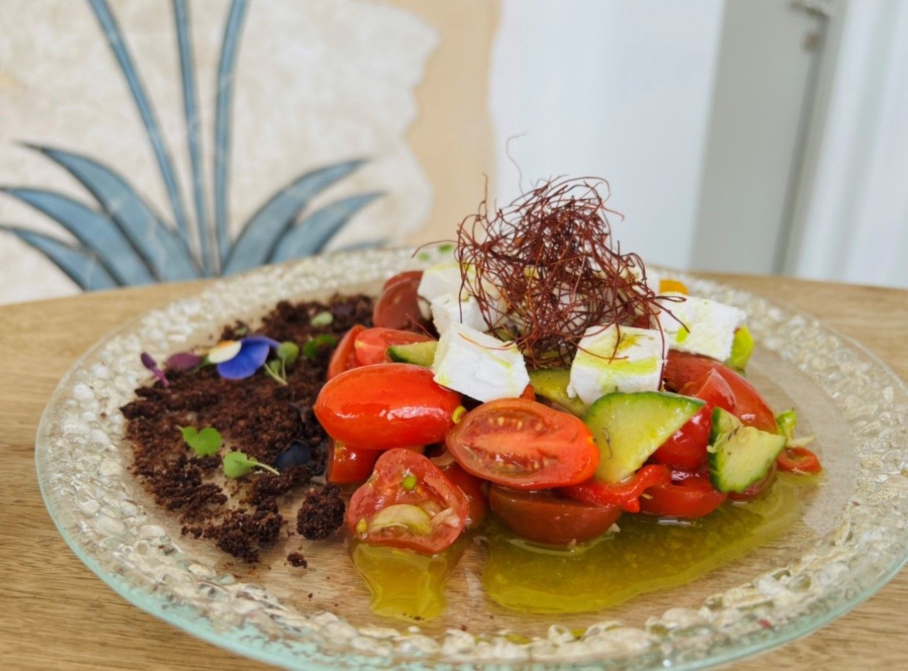 A glass plate with a salad consisting of tomatoes, avocado, and feta cheese, garnished with microgreens and a sprig of rosemary.