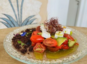 A glass plate with a salad consisting of tomatoes, avocado, and feta cheese, garnished with microgreens and a sprig of rosemary. - La Ponta Villas&suites (Santorini)