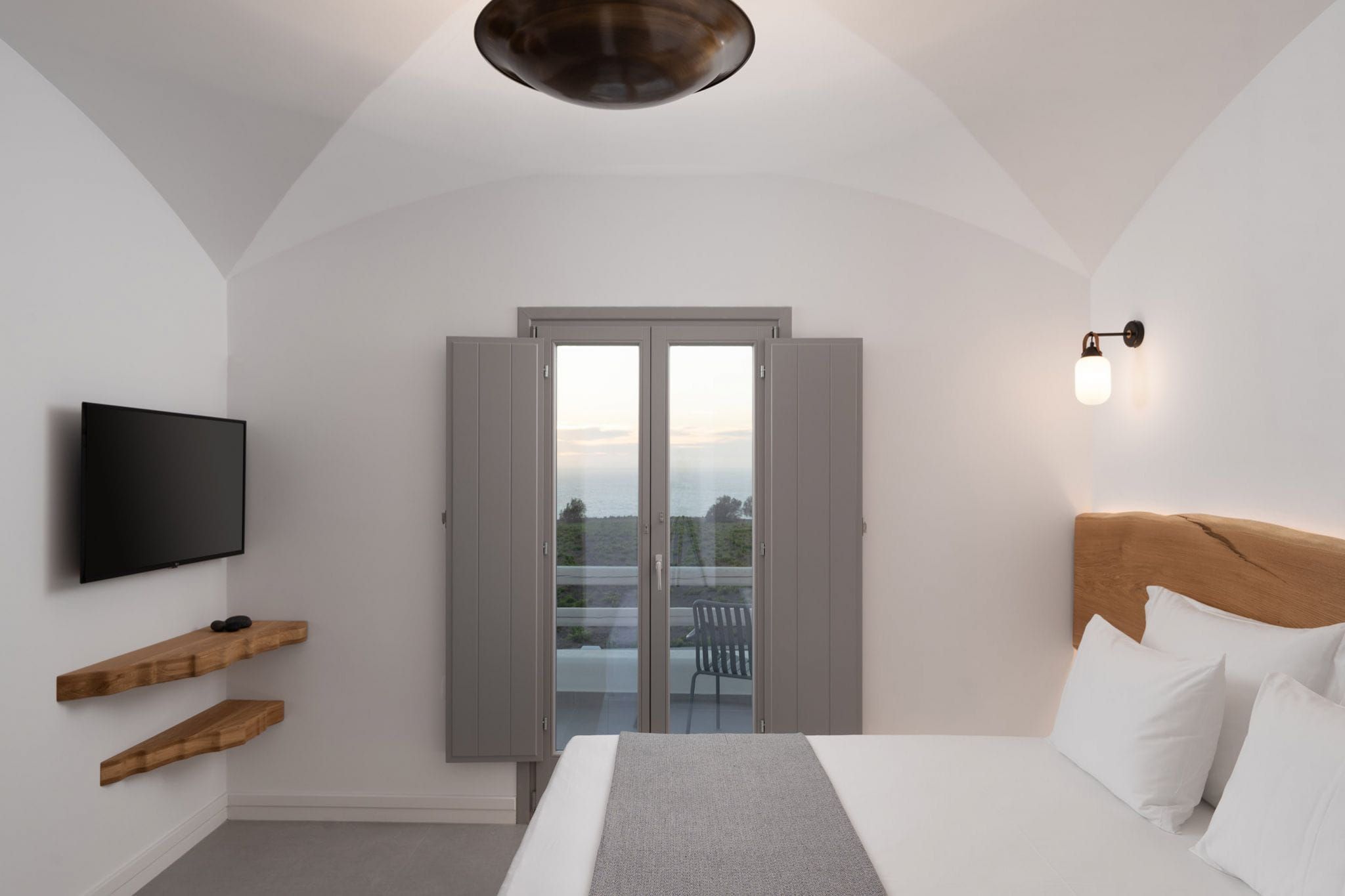 A bedroom with a bed, a TV, wooden shelves, a balcony door, and a wall-mounted light fixture.