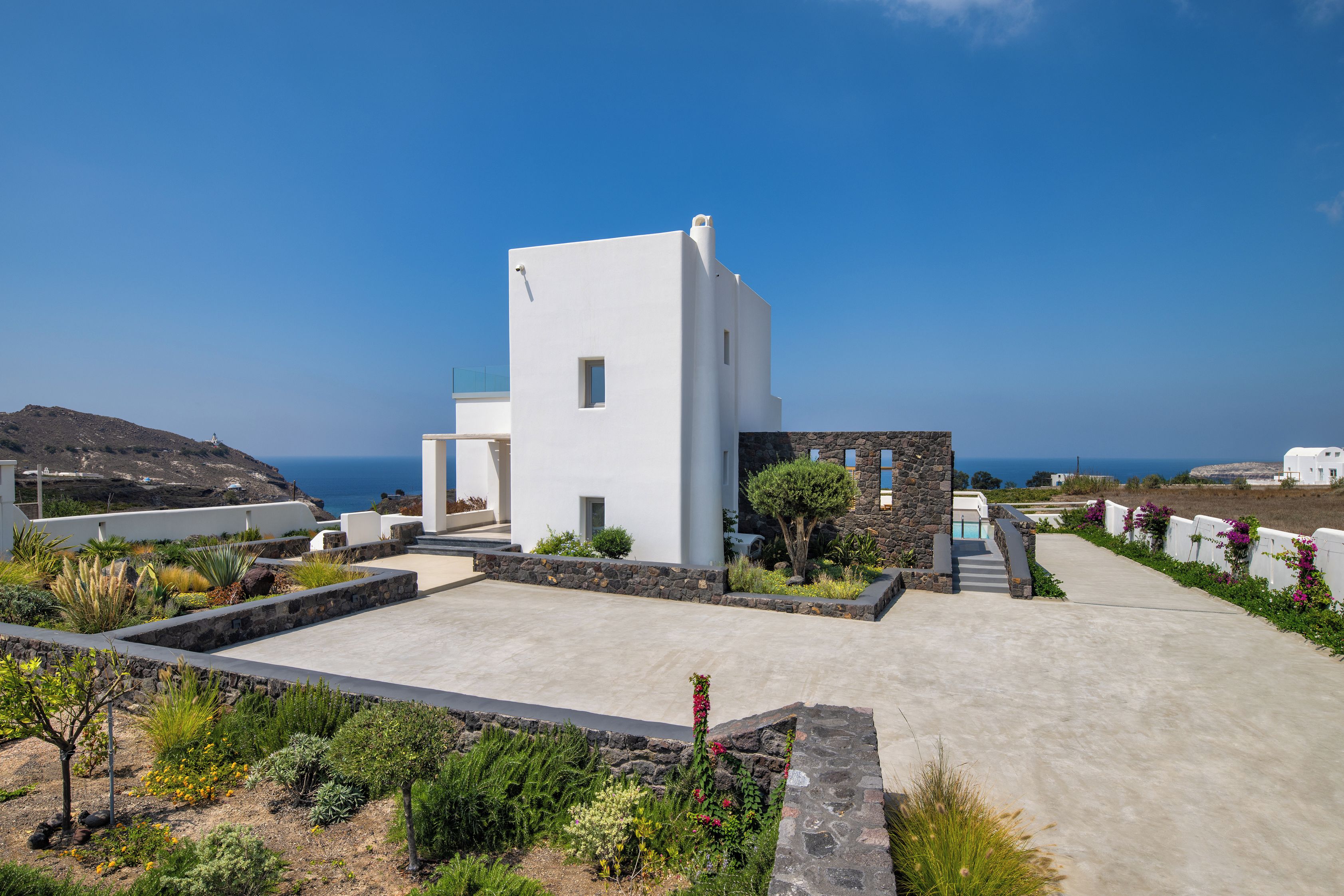 Um edifício moderno e branco, com detalhes em pedra, um pátio de concreto e vista para o mar.