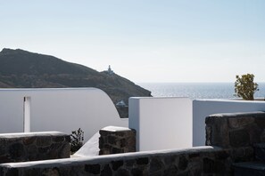A coastal view with a white building, stone walls, and a distant lighthouse. - La Ponta Villas&suites (Santorini)