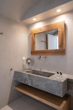 A bathroom with a wooden-framed mirror, a marble sink, and a small shelf. - La Ponta Villas&suites (Santorini)