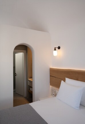A bedroom with a bed, a nightstand, a wall-mounted light, and an arched doorway leading to another room. - La Ponta Villas&suites (Santorini)