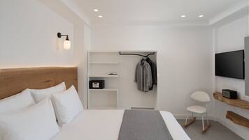 Una habitación de hotel moderna con cabecera de madera, cama con ropa de cama blanca, una manta gris, un televisor montado en la pared, perchero y una pequeña zona de estar con una silla.