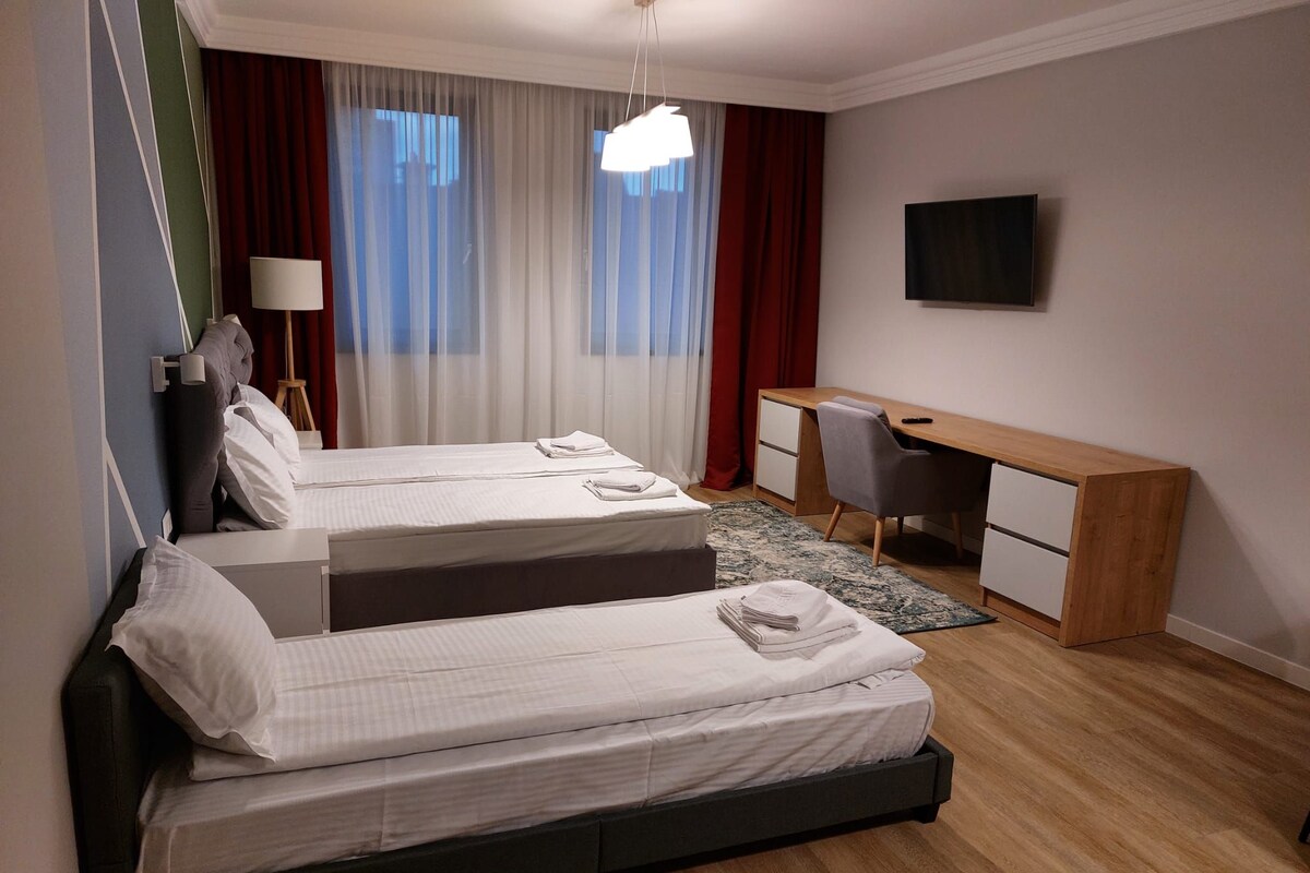A hotel room with two beds, a desk, a chair, a TV, and a window with curtains.