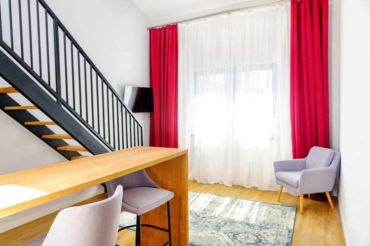 Un salon moderne avec une table en bois, deux chaises, un escalier, un téléviseur à écran plat et des rideaux rouges.