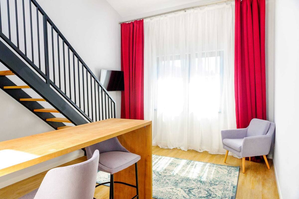 Un salon moderne avec une table en bois, deux chaises, un escalier, un téléviseur à écran plat et des rideaux rouges.