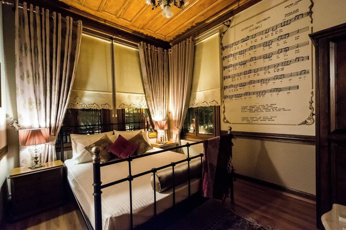 Una habitación con cama, cortinas, mesita de noche, lámpara, ventana y una pared con una partitura musical.