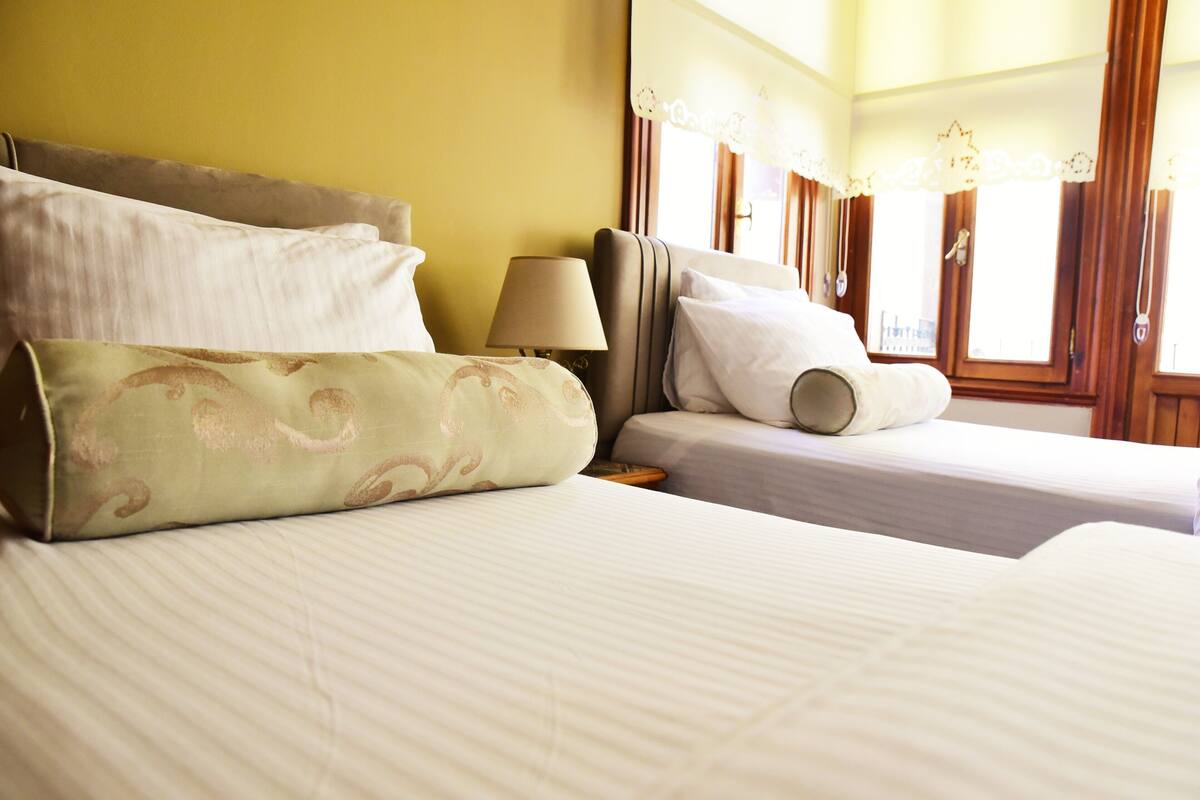 Habitación de hotel con dos camas, una mesita de noche, una lámpara y una ventana con cortinas.