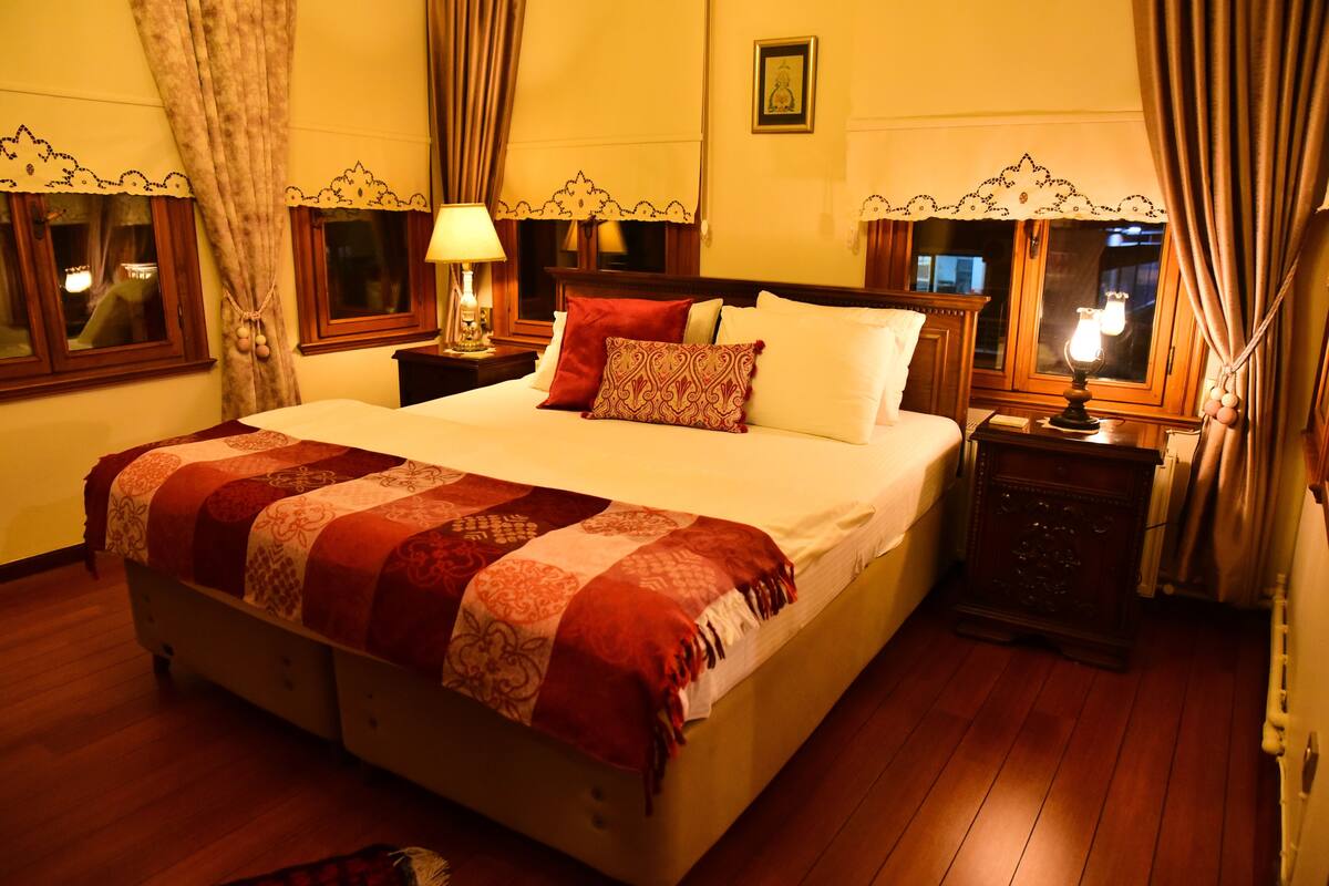 Habitación de hotel con una cama grande, dos mesitas de noche con lámparas, un aparador de madera y cortinas en las ventanas.