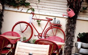 Un vélo rouge appuyé contre un mur, un arrosoir rouge, des chaises en osier et une plante en pot.