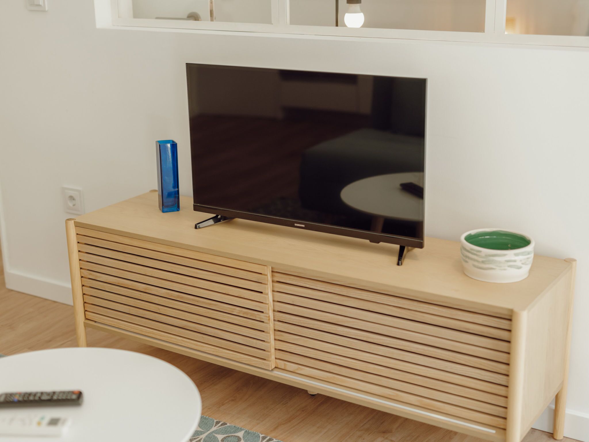 Televisão de tela plana sobre um console de madeira, com controle remoto e um objeto decorativo ao lado.