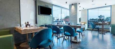 A modern dining area with wooden tables, blue chairs, and large windows offering a view of the city.