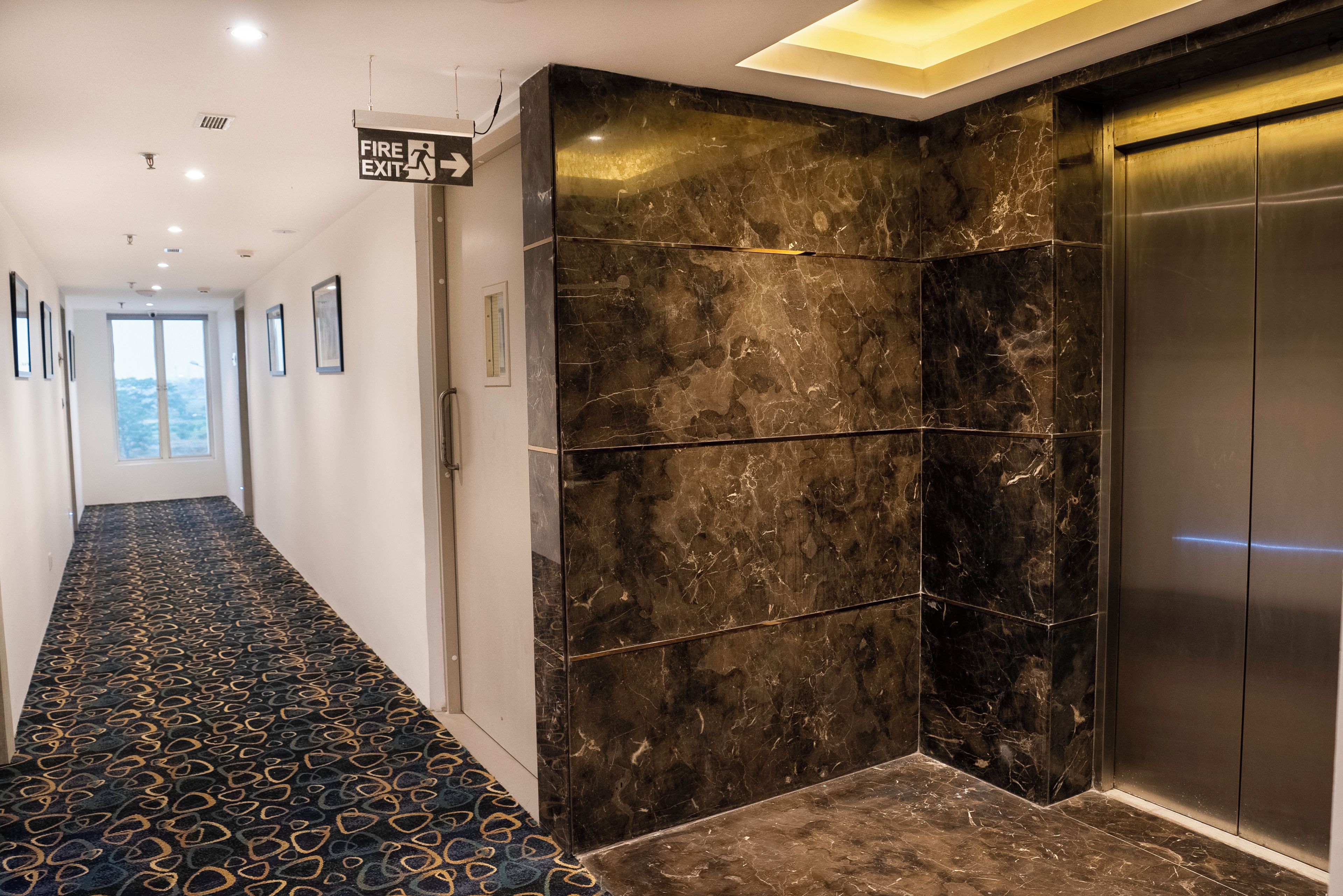 A hallway with a patterned carpet, a fire exit sign, a marble wall, and an elevator.