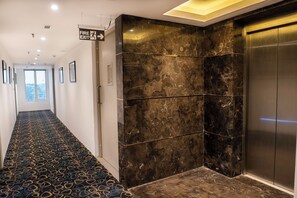 A hallway with a patterned carpet, a fire exit sign, a marble wall, and an elevator. - Poiisse - A Boutique Hotel (Kolkata)