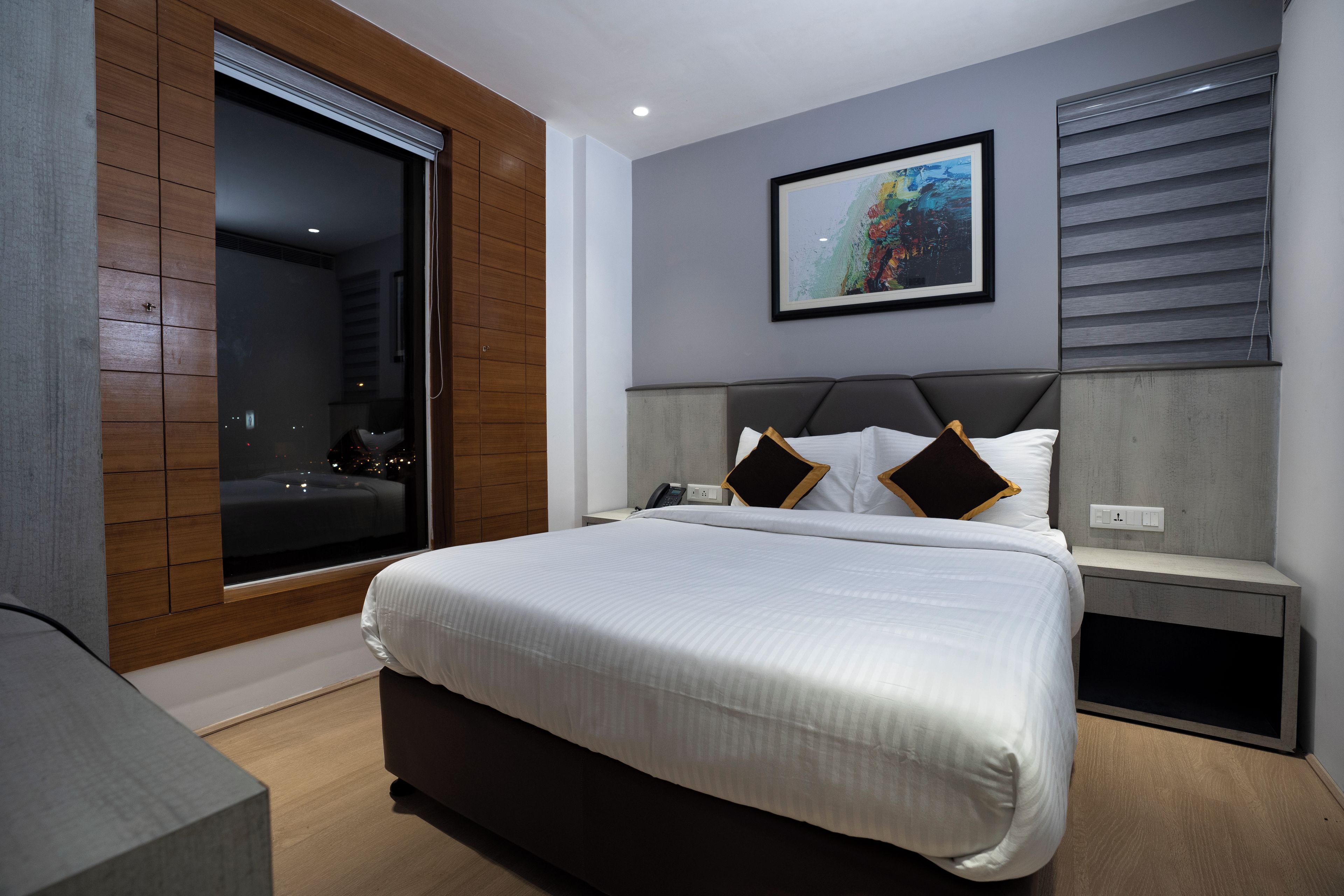 A modern bedroom with a large bed, a bedside table, a nightstand, a window with blinds, and a framed picture on the wall.