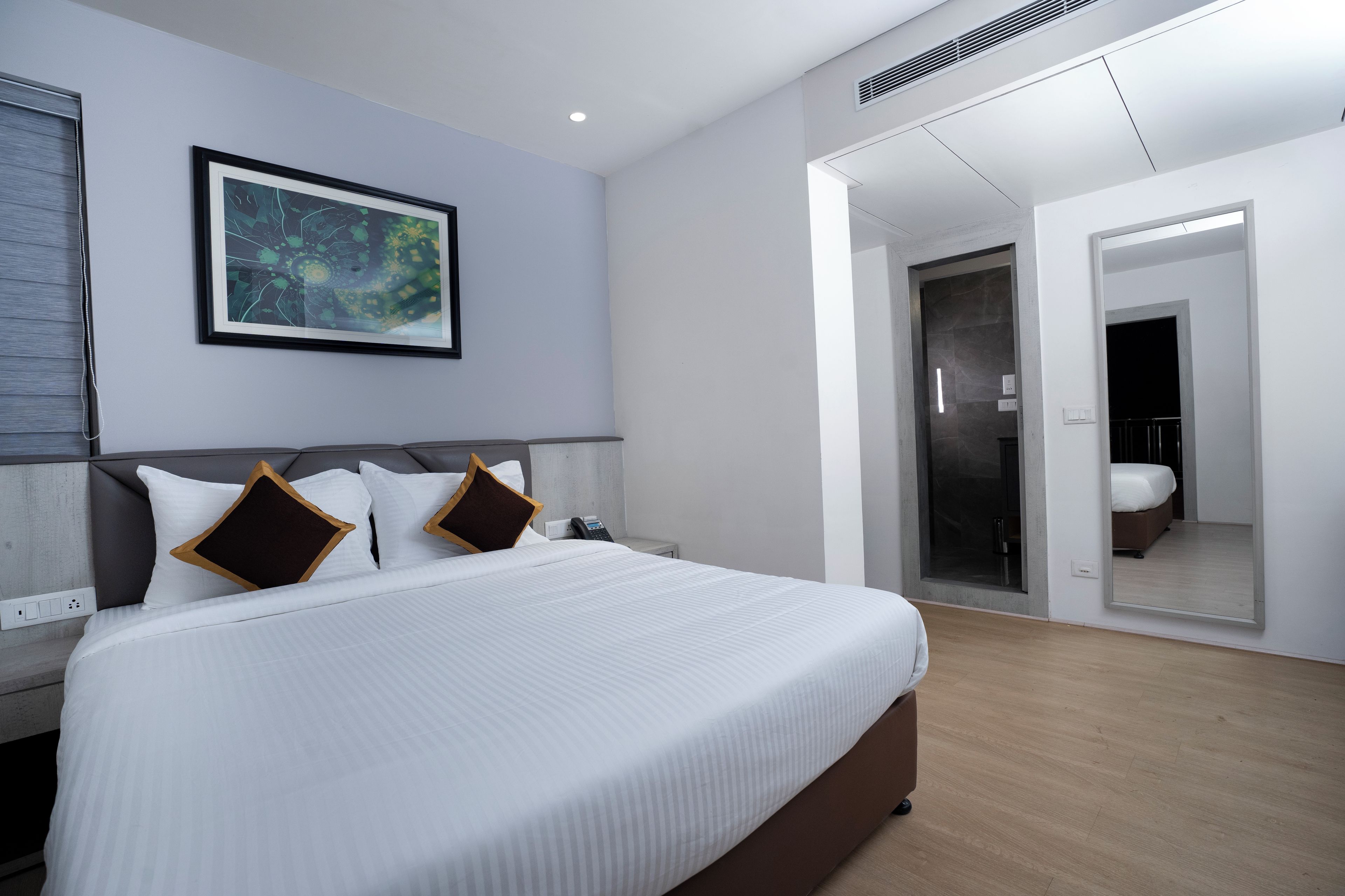 A modern hotel room with a large bed, a framed artwork on the wall, a walk-in closet, and a bathroom visible through a glass door.