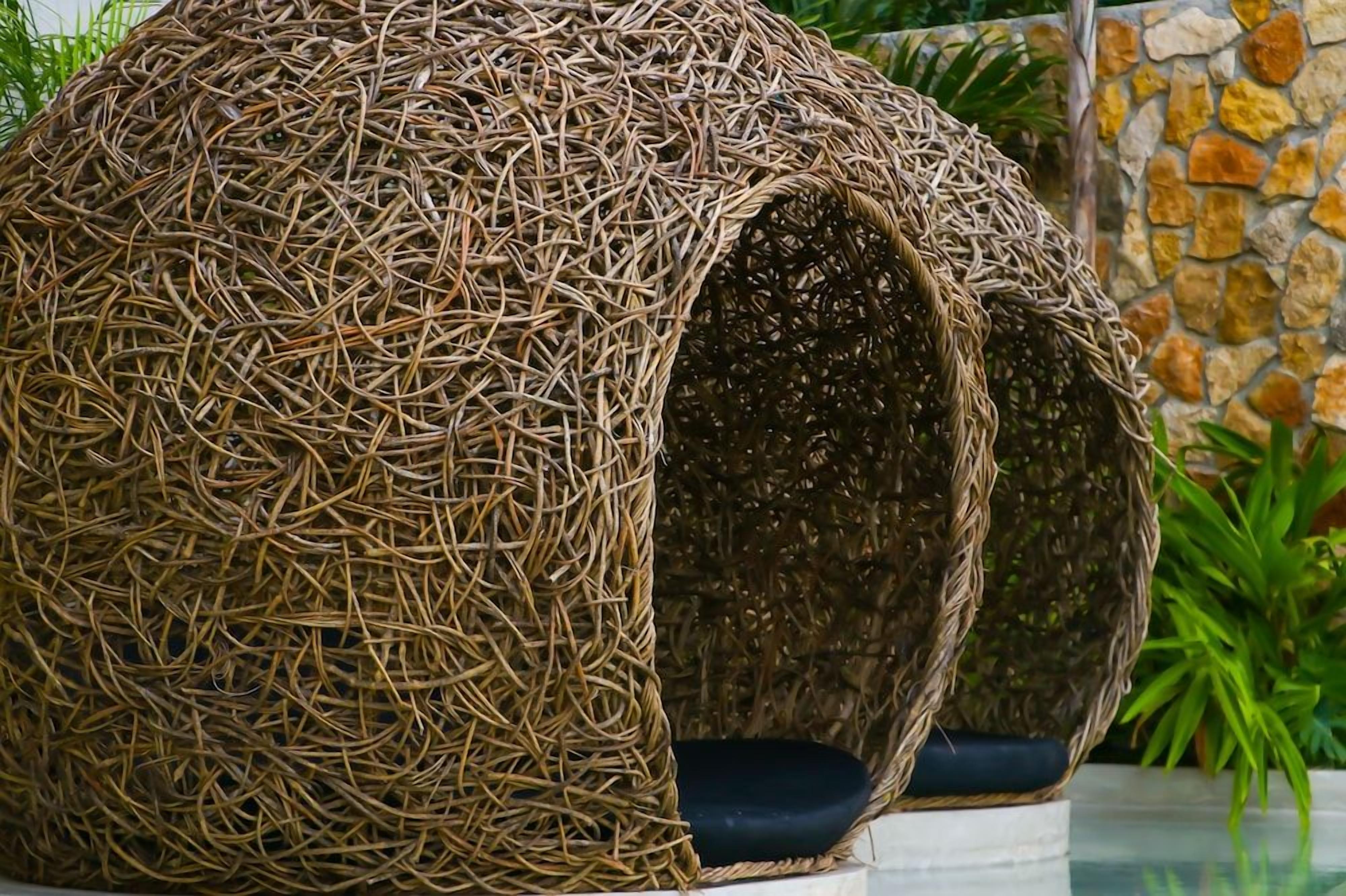 Una estructura esférica de mimbre con un cojín en su interior, ubicada frente a un muro de piedra y follaje.