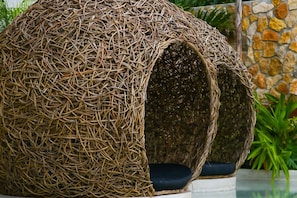 A spherical wicker structure with a cushion inside, set against a stone wall and greenery.
