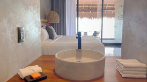 A hotel room with a large round sink, a bed with white bedding, a bedside lamp, and a view of the beach through the window.