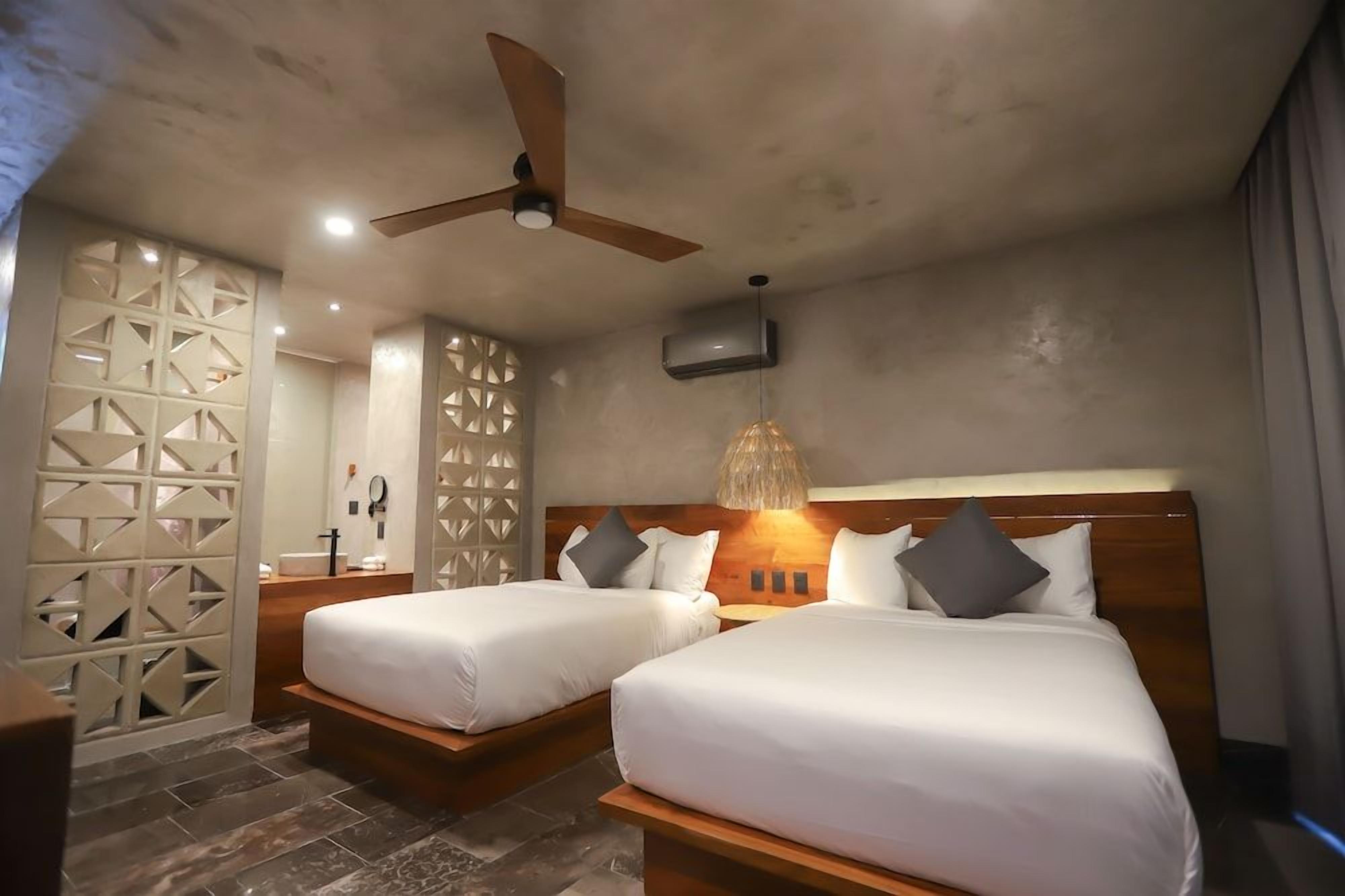 Habitación de hotel con dos camas, ventilador de techo, televisor montado en la pared y una lámpara decorativa.