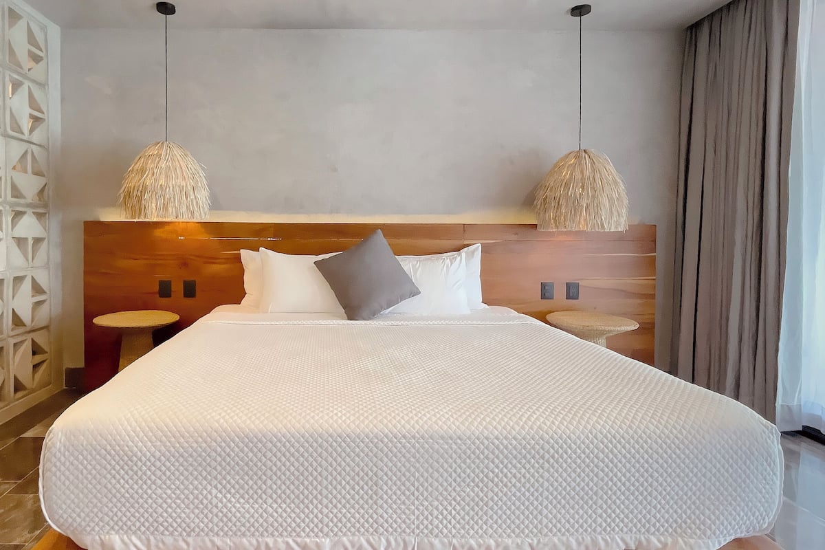 Uma cama grande com edredom branco acolchoado, duas mesas de cabeceira, uma cabeceira de madeira e duas luminárias pendentes.