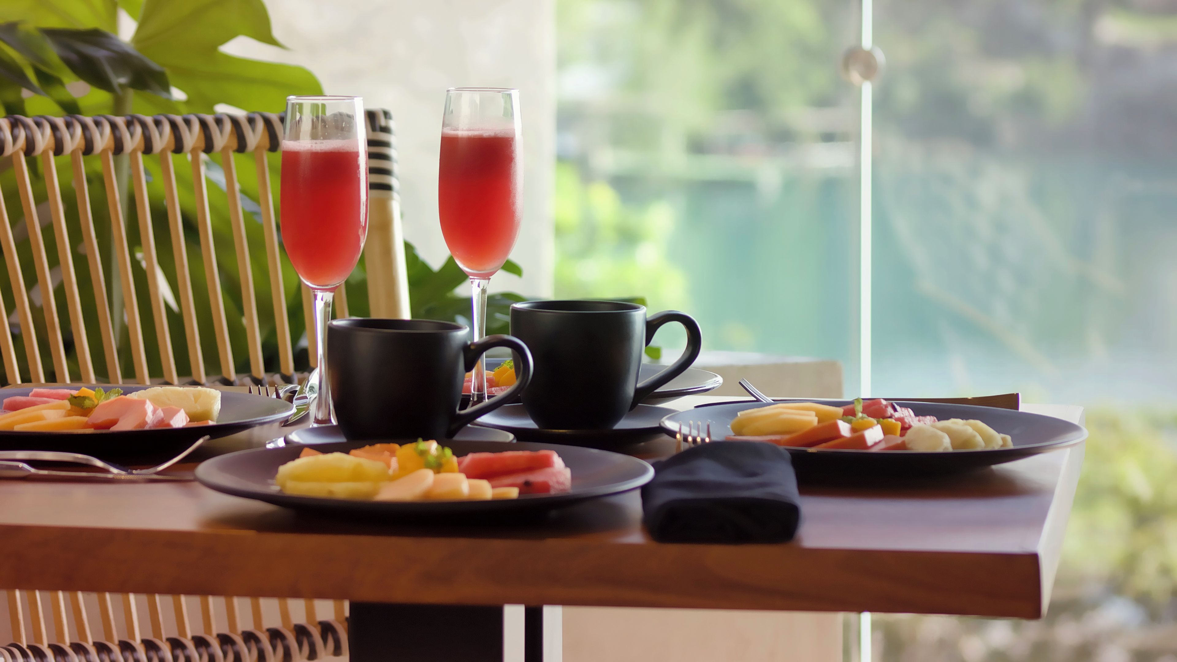 A breakfast setting with plates of fruit, two glasses of red beverage, and black cups on a table with a view of greenery.