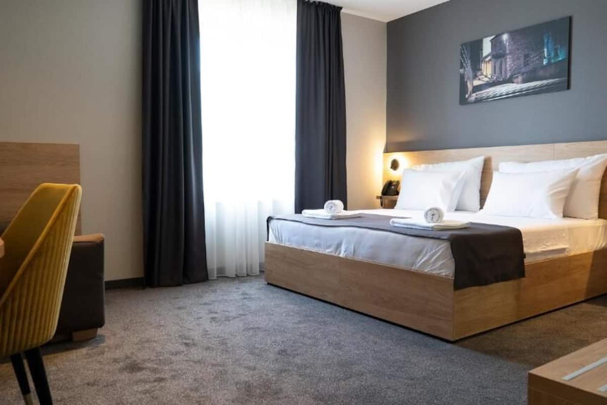 Quarto de hotel com cama, escrivaninha, cadeira e uma grande janela com cortinas.