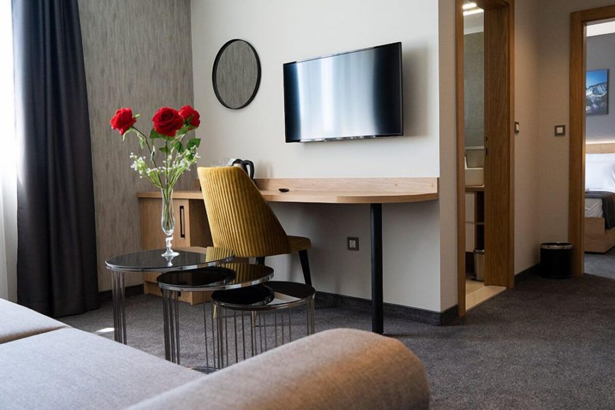 A hotel room with a bed, a desk with a chair, a TV, a vase with flowers, and a view of another room.