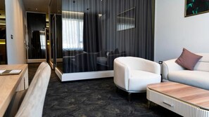 A modern hotel room with a glass partition, a white armchair, a sofa, a wooden coffee table, and a small desk with a chair. - Ha Hotel (Mostar)