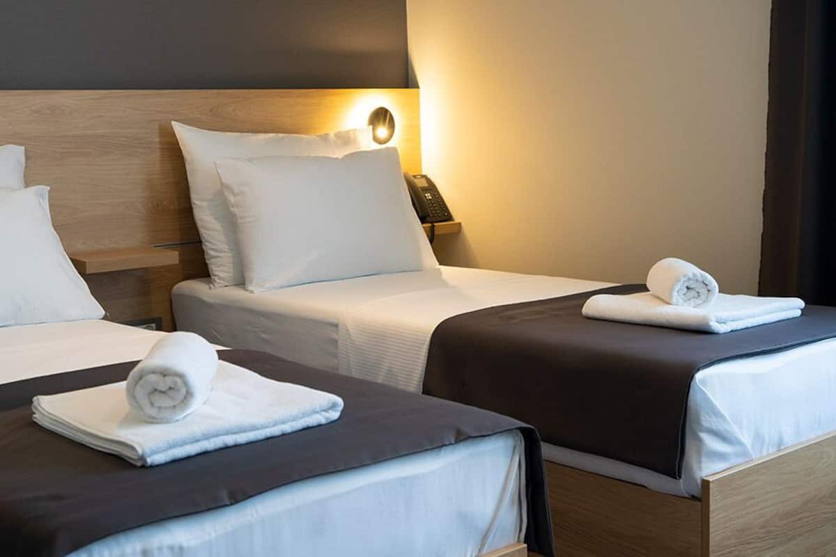 Quarto de hotel com duas camas, cada uma com um travesseiro, uma mesa de cabeceira com abajur e uma toalha enrolada na cama.