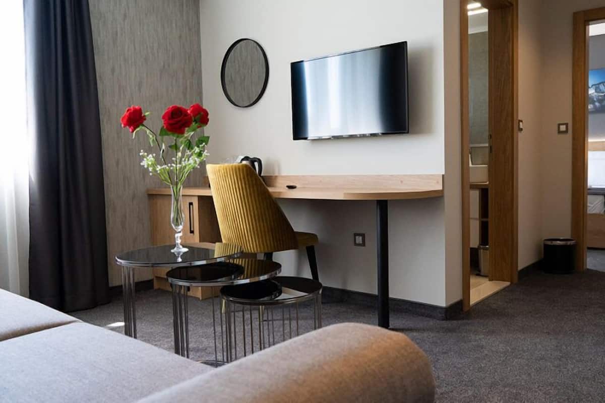 Quarto de hotel com cama, escrivaninha com cadeira, televisão, vaso com flores e vista para outro quarto.