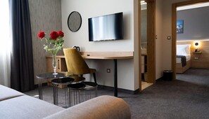 Une chambre d’hôtel avec un lit, un bureau et une chaise, une télévision, un vase de fleurs et une vue sur une autre chambre.