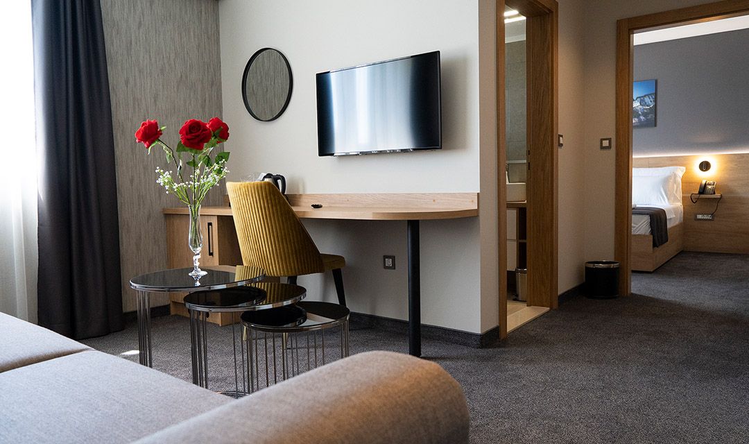 Habitación de hotel con cama, televisor montado en la pared, una pequeña mesa con sillas, un florero con flores y un escritorio de madera.