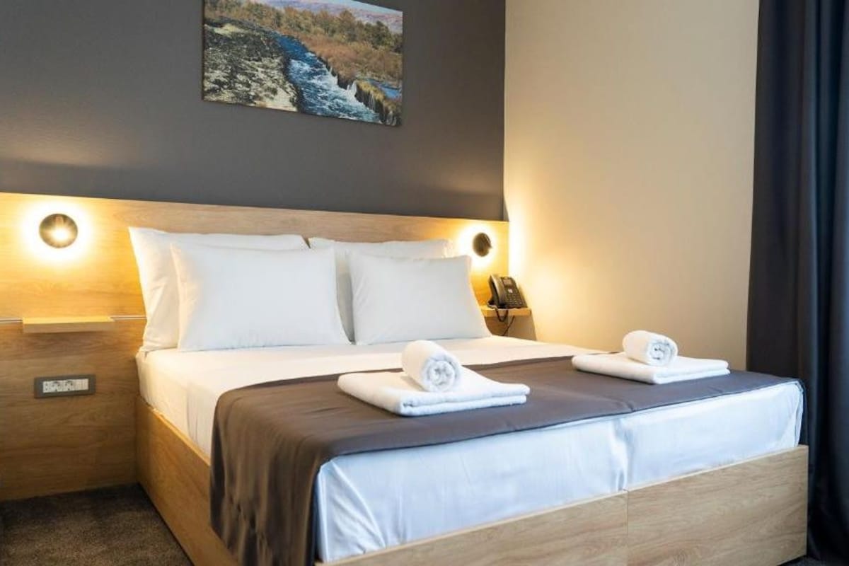 A hotel room with a bed, two bedside tables, a painting on the wall, and a window with curtains.
