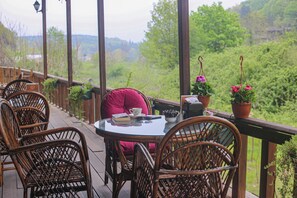 A covered outdoor seating area with wicker chairs and a table, surrounded by potted plants and overlooking a lush green landscape.
